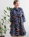 Woman wearing a patterned dress standing next to a plant indoors.